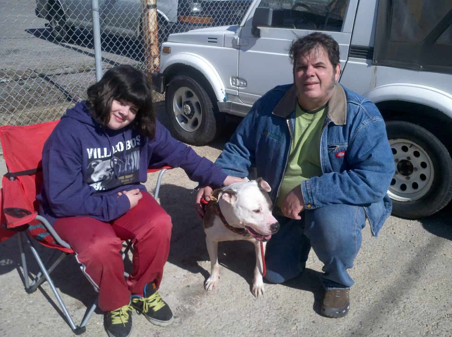A person in a red tracksuit sits in a folding chair next to another person in a blue jacket; both are petting a white dog. They are outdoors by a fence with parked vehicles in the background.