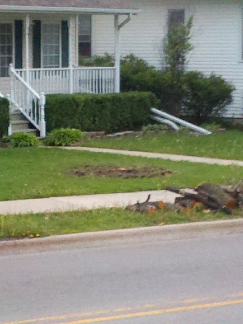 Tree #4 REVISED A broken metal railing lies in the grass near a sidewalk in front of a house with a porch, indicating recent damage or an accident.