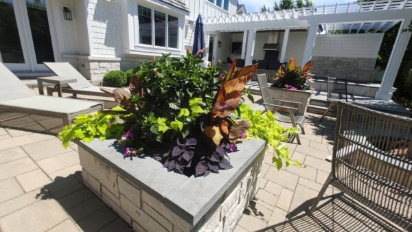 A large stone planter filled with green, purple, and orange foliage sits on a sunny patio near lounge chairs, outdoor dining furniture, and a grill area outside a white house.