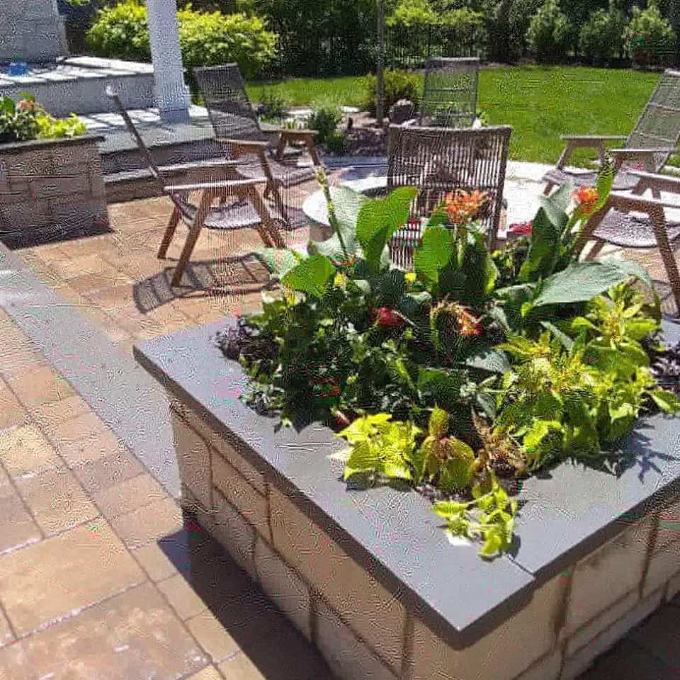 A square stone planter filled with green and flowering plants sits on a sunny patio with wooden chairs and a lawn in the background.
