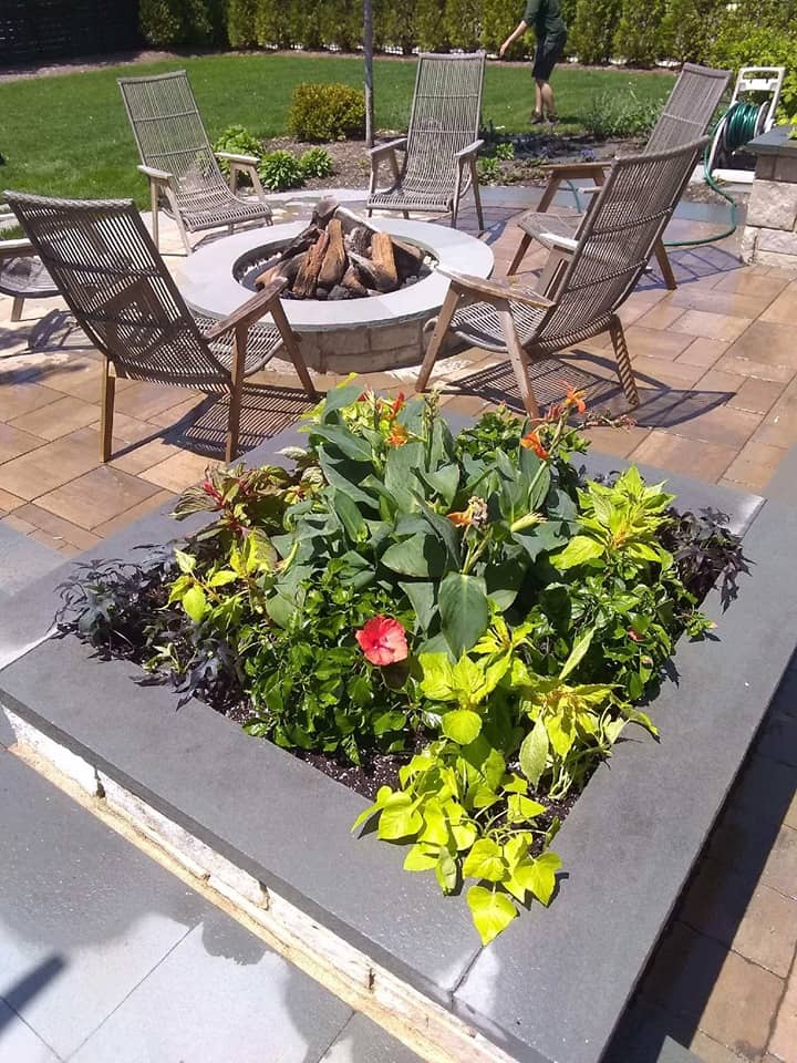 Raised garden bed with colorful flowers and leafy plants in the foreground, surrounded by a stone patio with wooden chairs arranged around a fire pit; a green lawn is visible in the background.