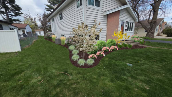 A landscaped yard with green grass, flowering trees, and colorful flower beds surrounds the corner of a beige house with white trim. A pathway leads to the front steps.