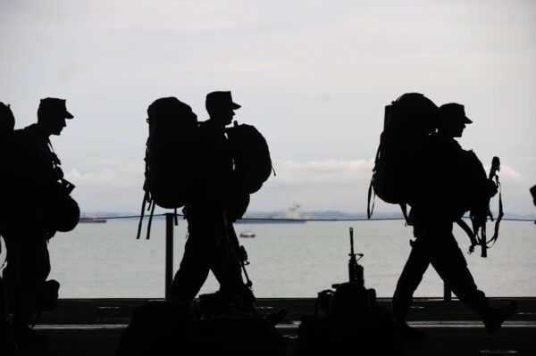 Silhouettes of soldiers carrying large backpacks walk beside a body of water, with ships visible in the distance and a cloudy sky overhead.