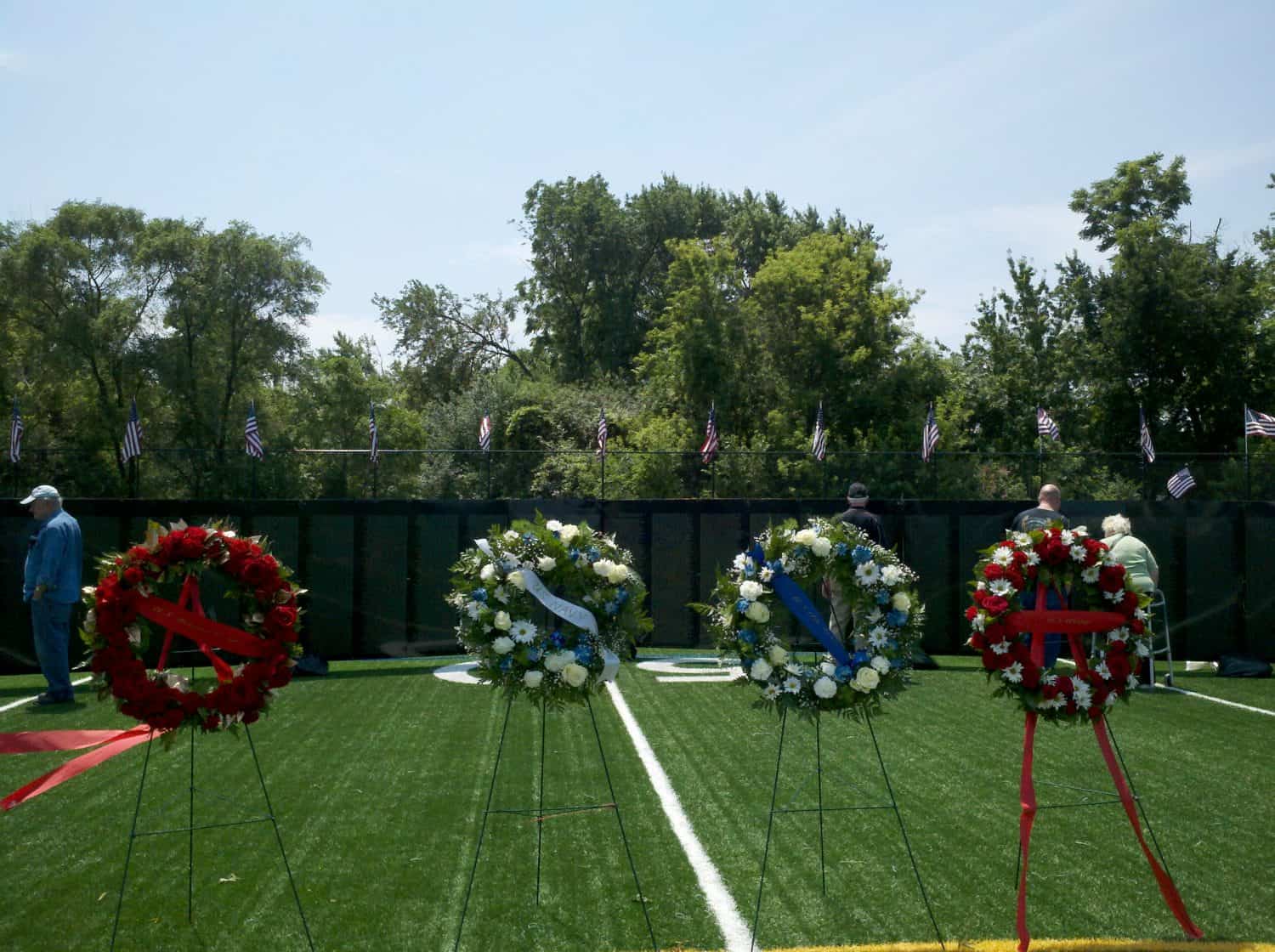 Four floral wreaths on stands are displayed on a sports field, with people standing near a black memorial wall in the background. Small American flags are lined along the top of the wall. Trees border the scene.