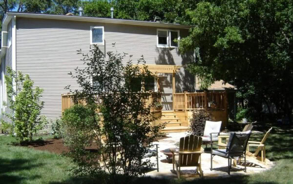 A backyard features a wooden deck attached to a house, outdoor chairs surrounding a small fire pit on a stone patio, and various trees and shrubs providing shade.