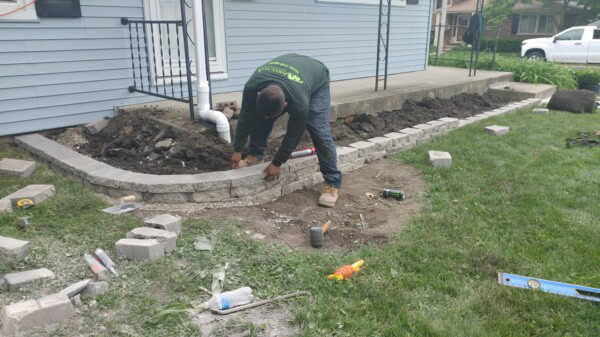 A person bends over to build a stone retaining wall along a house. Tools, bricks, and supplies are scattered on the ground beside them. The wall construction is partially completed.