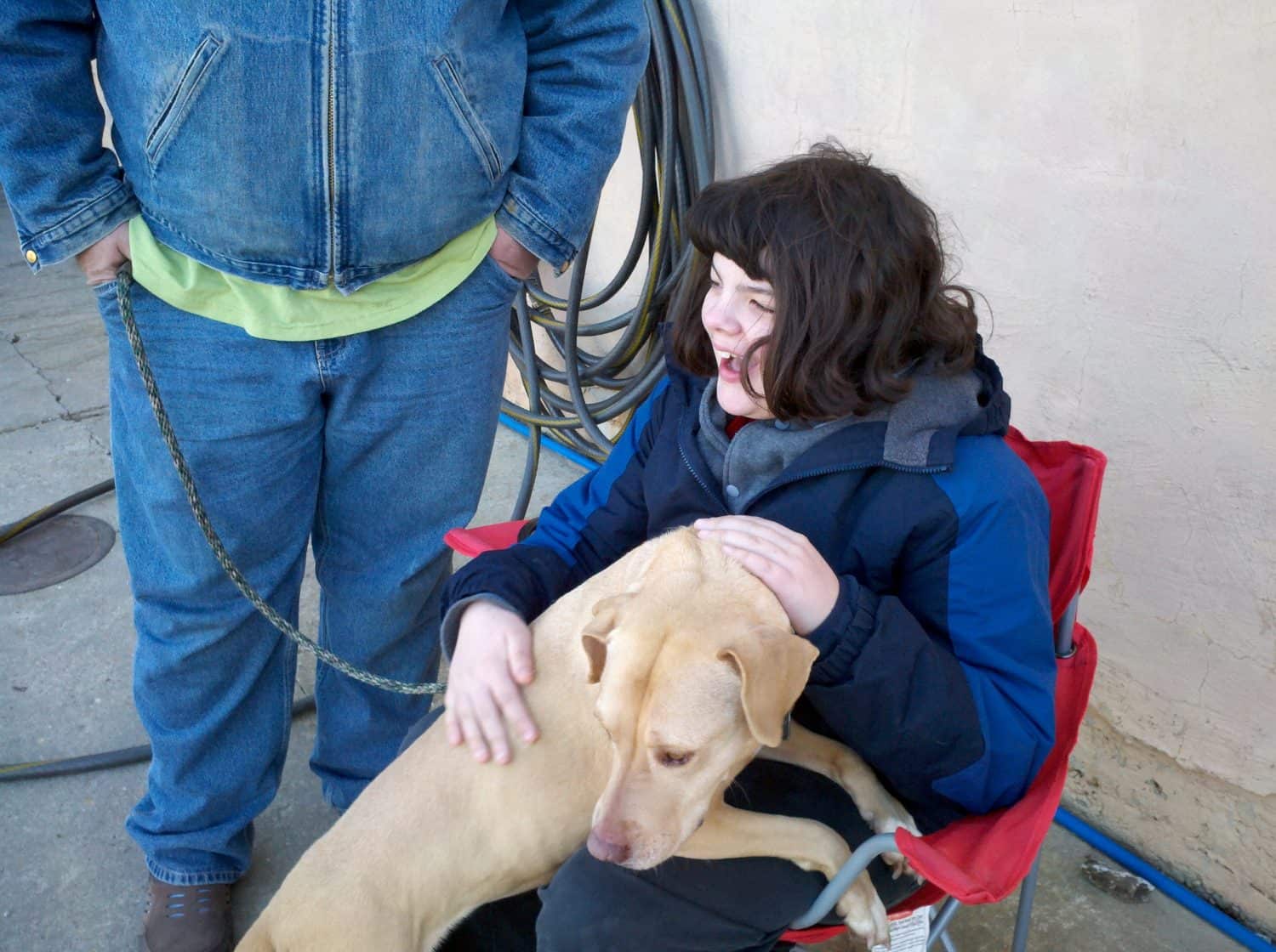 A person sitting in a red chair smiles and pets a tan dog in their lap. Another person stands nearby, holding the dogs leash. A coiled hose hangs on the wall behind them.