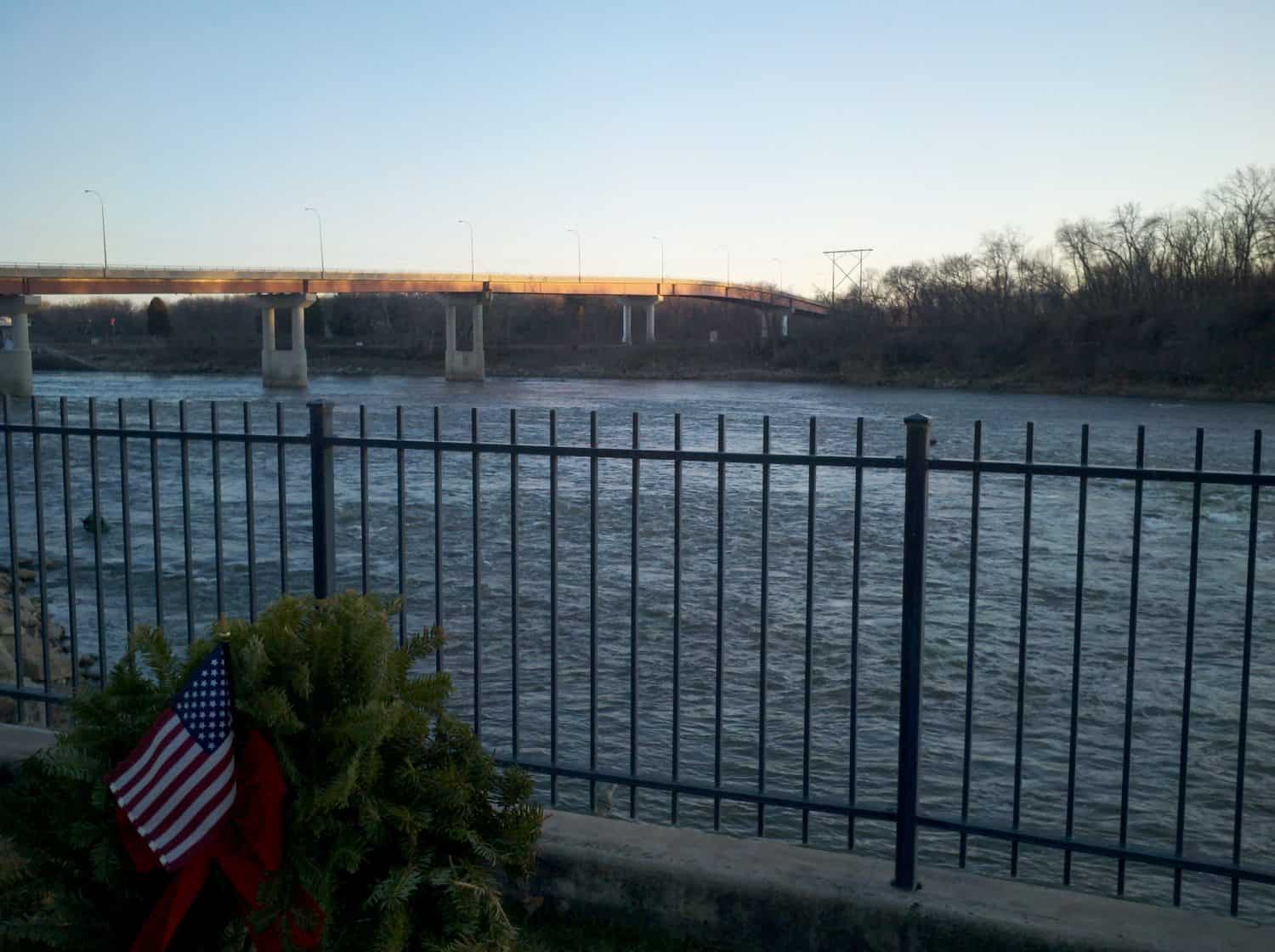 A metal fence overlooks a wide river with a bridge in the background. In the foreground, a wreath with an American flag is attached to the fence. Trees line the far bank under a clear sky.