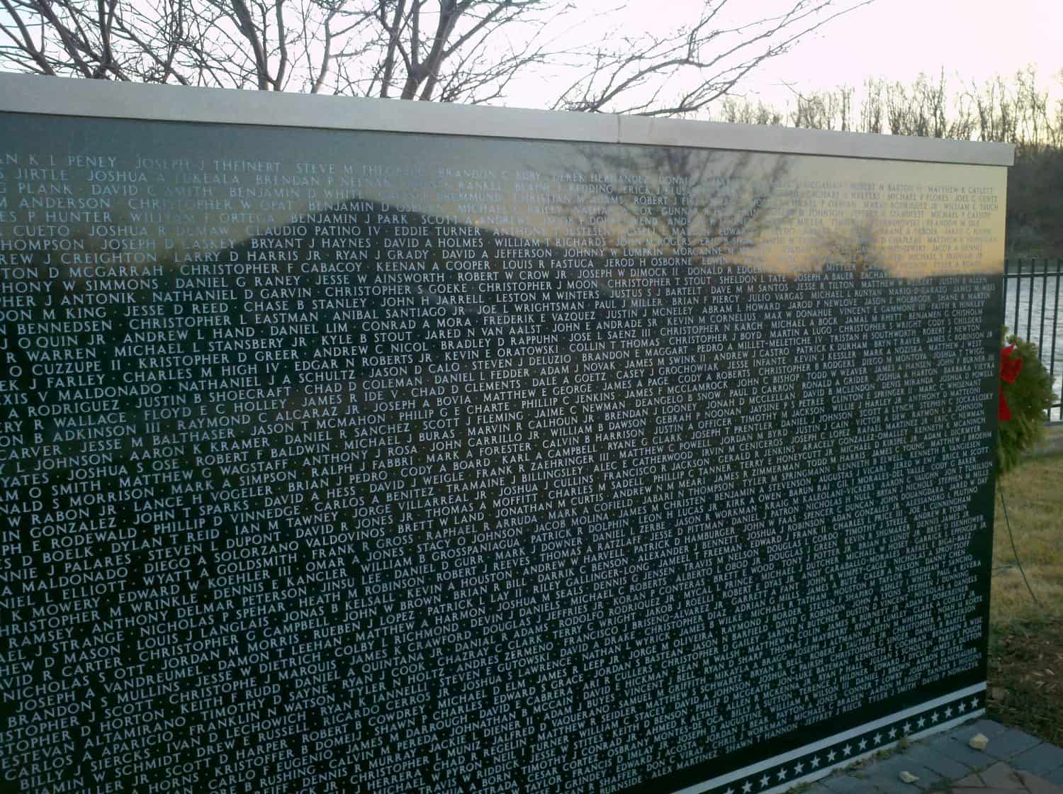 A black granite memorial wall engraved with many names, outdoors by a river and leafless trees, with a wreath visible at one end.