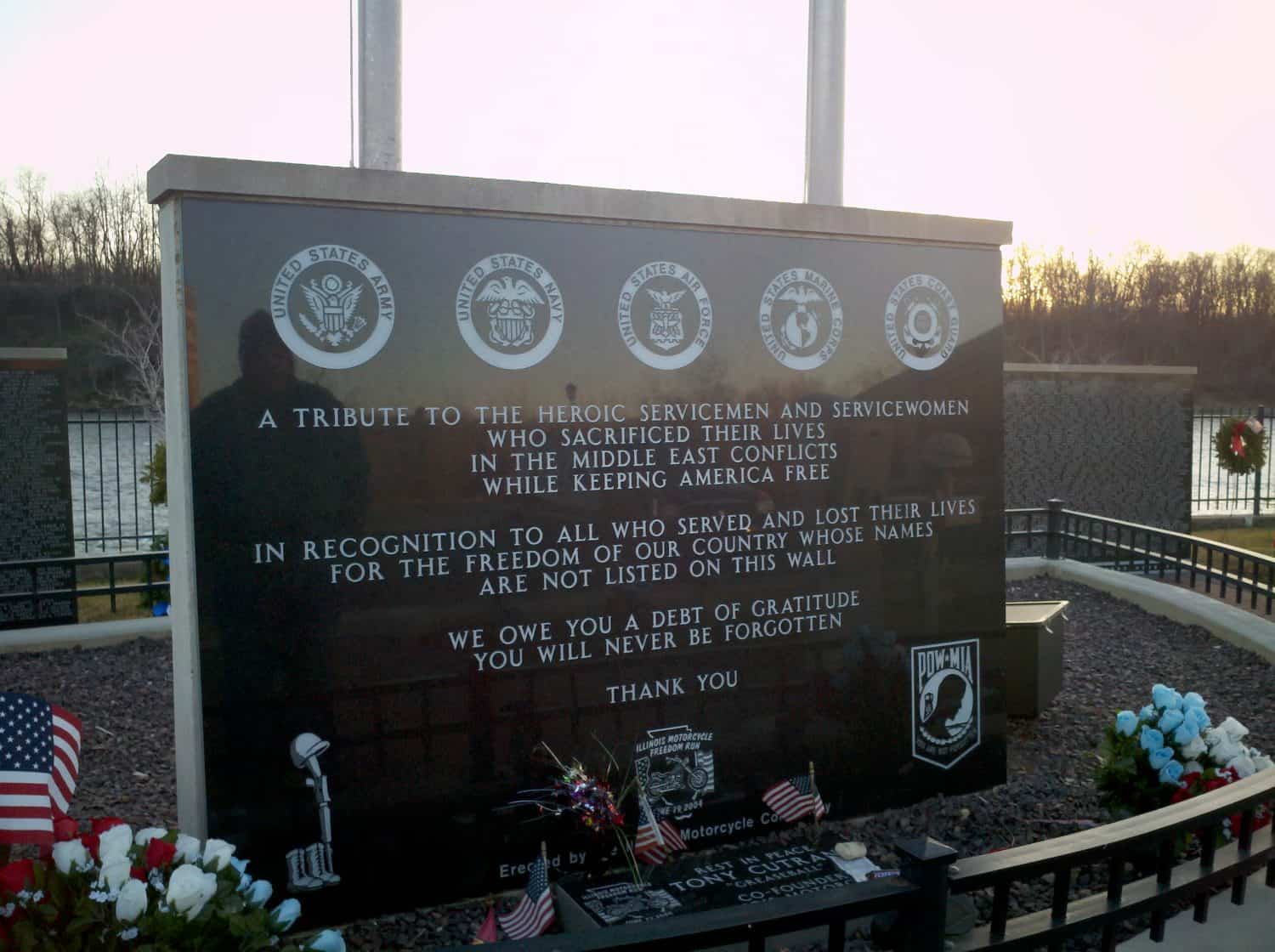 A memorial wall honors servicemen and servicewomen who died in Middle East conflicts. Flags, wreaths, and military emblems are displayed. The text expresses gratitude for their sacrifice.