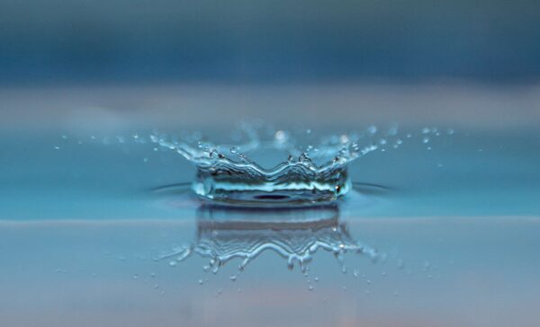 A close-up of a water droplet splashing into a calm blue surface, creating a crown-shaped ripple and reflection.