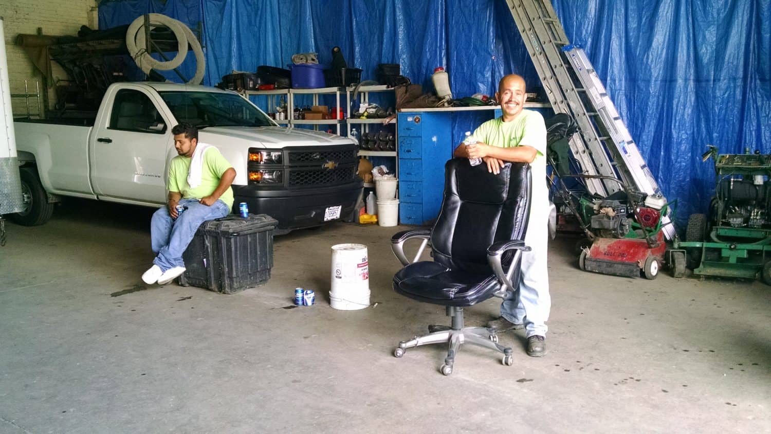 Two men in a garage with tools, a truck, and lawn equipment. One man sits on a black box, drinking from a can, while the other stands, smiling, with his hands on an office chair. Blue tarp hangs in the background.