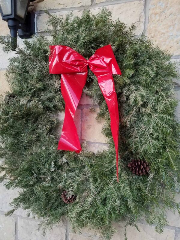 A green pine wreath with two pinecones and a large red bow hangs on a stone wall near an outdoor light fixture.