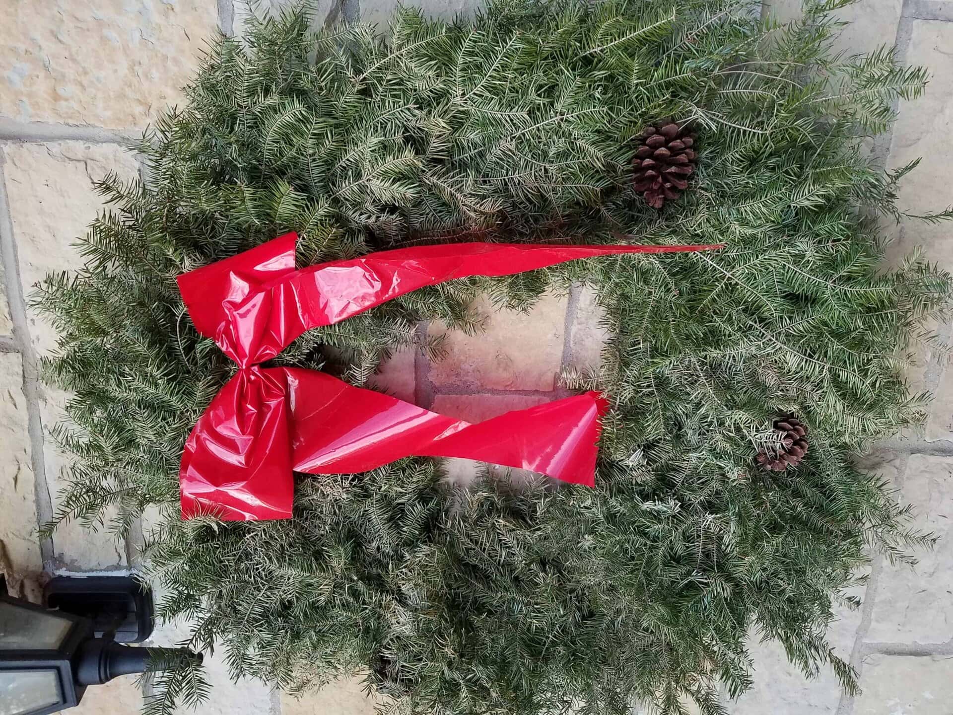A green holiday wreath with a large red bow and two pinecones hangs on a stone wall.